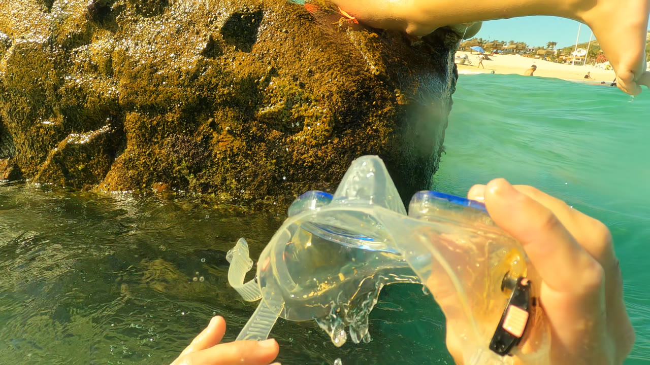 Hand holding a clear snorkel mask near rocks and ocean.