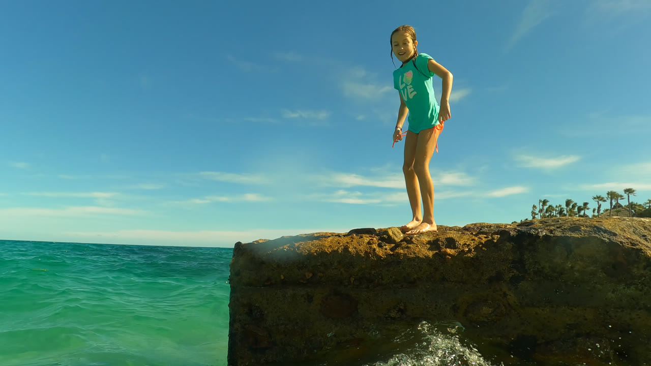 The girl leans slightly forward on the rock above clear green-blue water, with the horizon line and low coastal buildings visible in the background.