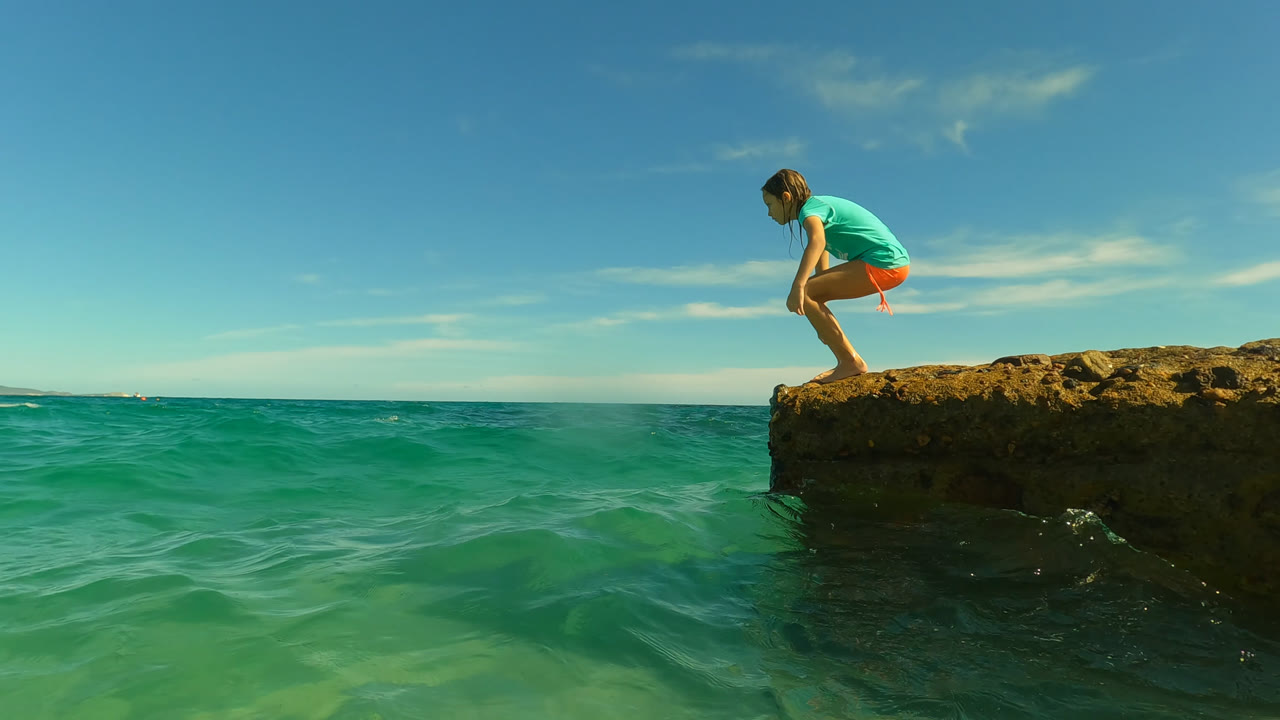 At San Jose Del Cabo, Mexico, a young girl crouches on the edge of a rocky outcrop above clear green-blue ocean under a bright sky.