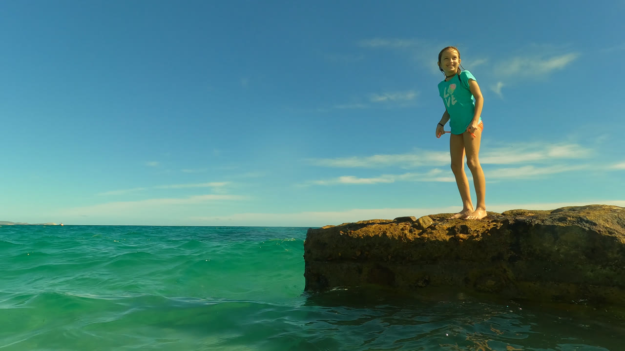 Young woman standing on a rocky outcrop above turquoise ocean water.