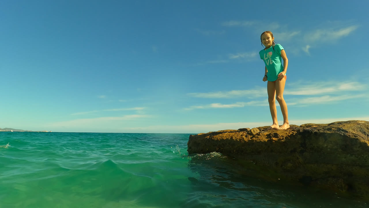 At San Jose Del Cabo, Mexico, the young woman remains centered on the rock while green-blue water ripples across the foreground under bright sun.