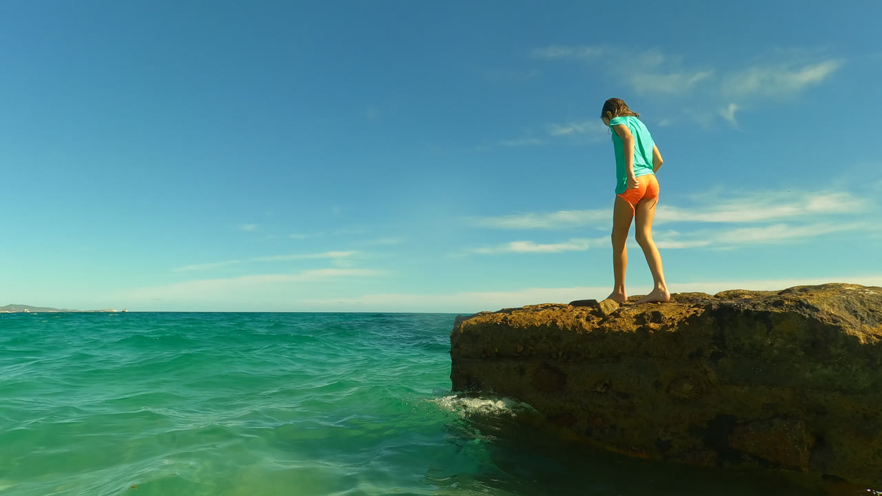 At 0:00 in San Jose Del Cabo, Mexico, a young girl stands near the left side of a rock ledge above turquoise water, looking down with the horizon behind her.
