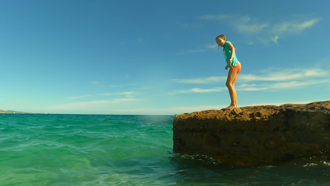 At 0:01 in San Jose Del Cabo, Mexico, the girl leans slightly forward on the rough rock edge while green-blue water rises against the ledge below.
