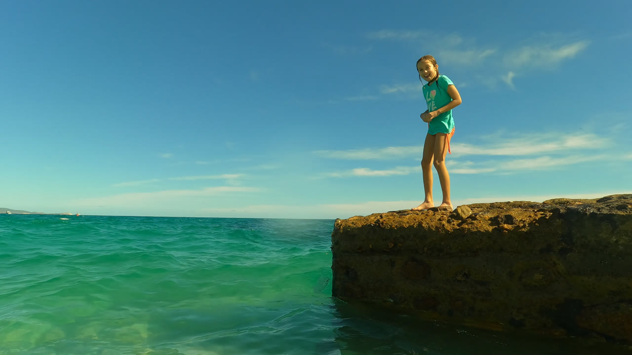 Young woman standing barefoot on a rocky ledge above turquoise ocean water.