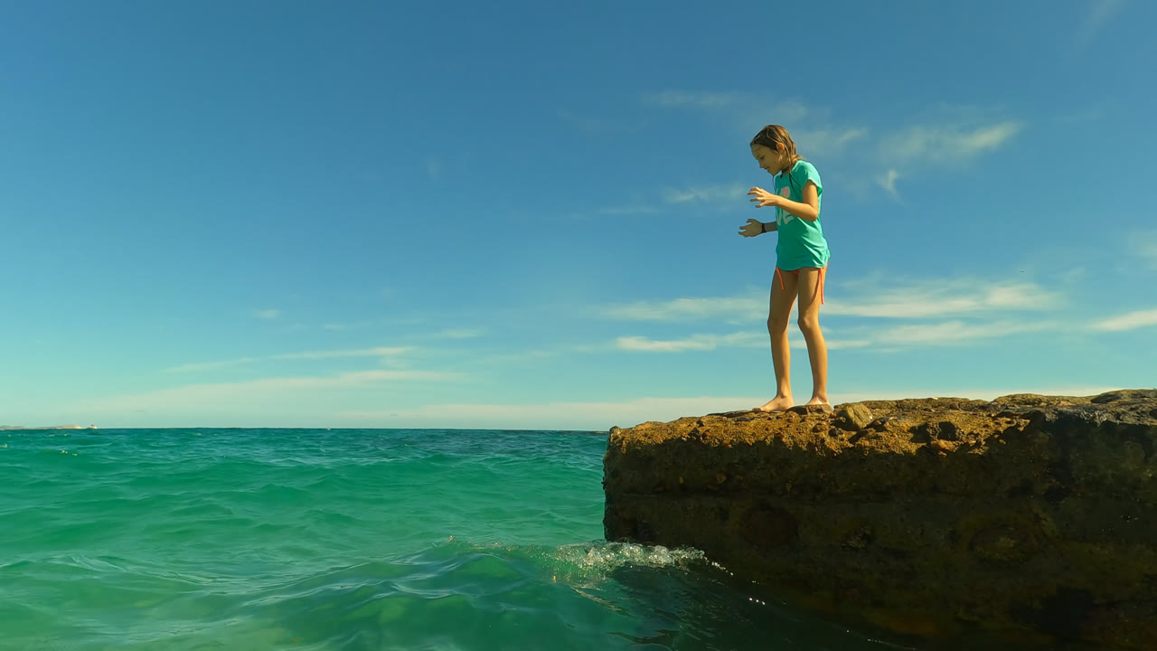 Young woman looks down from the rocky ledge toward the turquoise water in San Jose Del Cabo, Mexico, with open sea and a flat horizon behind her.