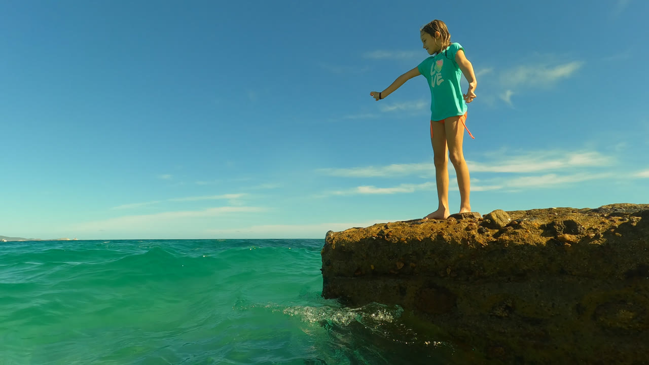 Young girl in a turquoise shirt stands near the edge of a rocky outcrop above green-blue water in San Jose Del Cabo, Mexico, looking down toward the ocean.