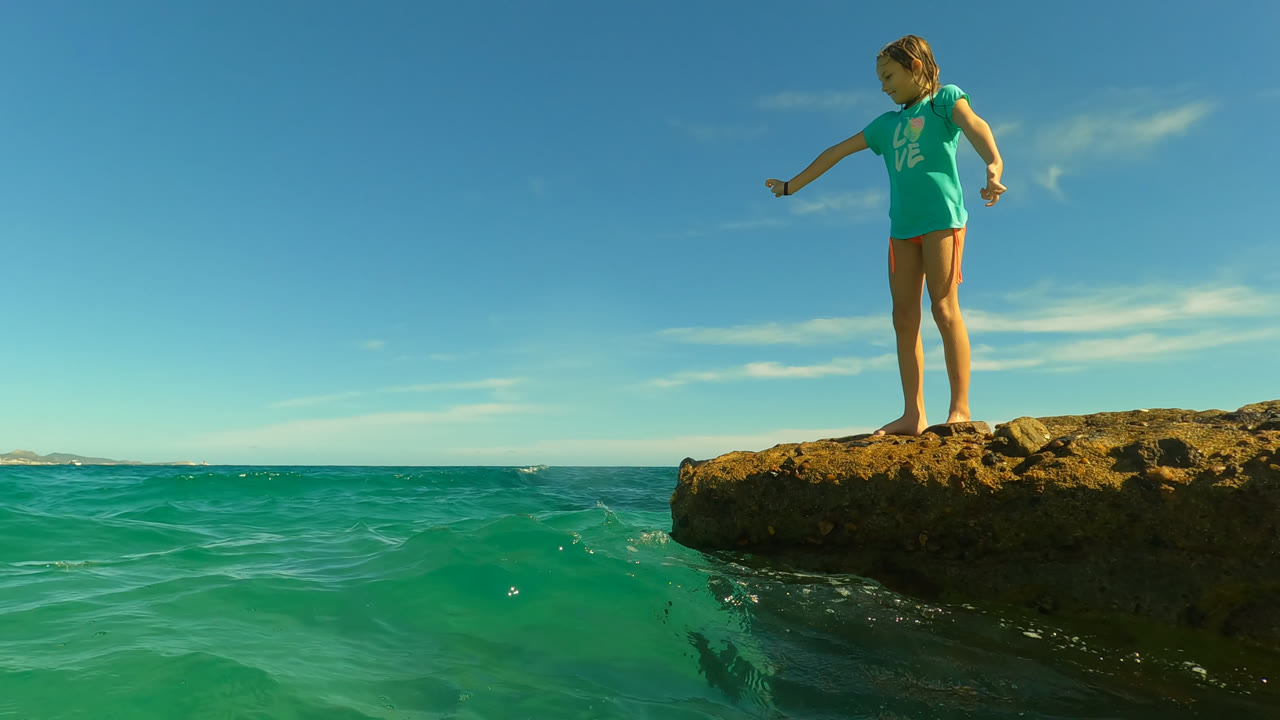 Girl stands barefoot on the uneven rock ledge in San Jose Del Cabo, Mexico, with small waves breaking below and a low strip of distant land on the horizon.