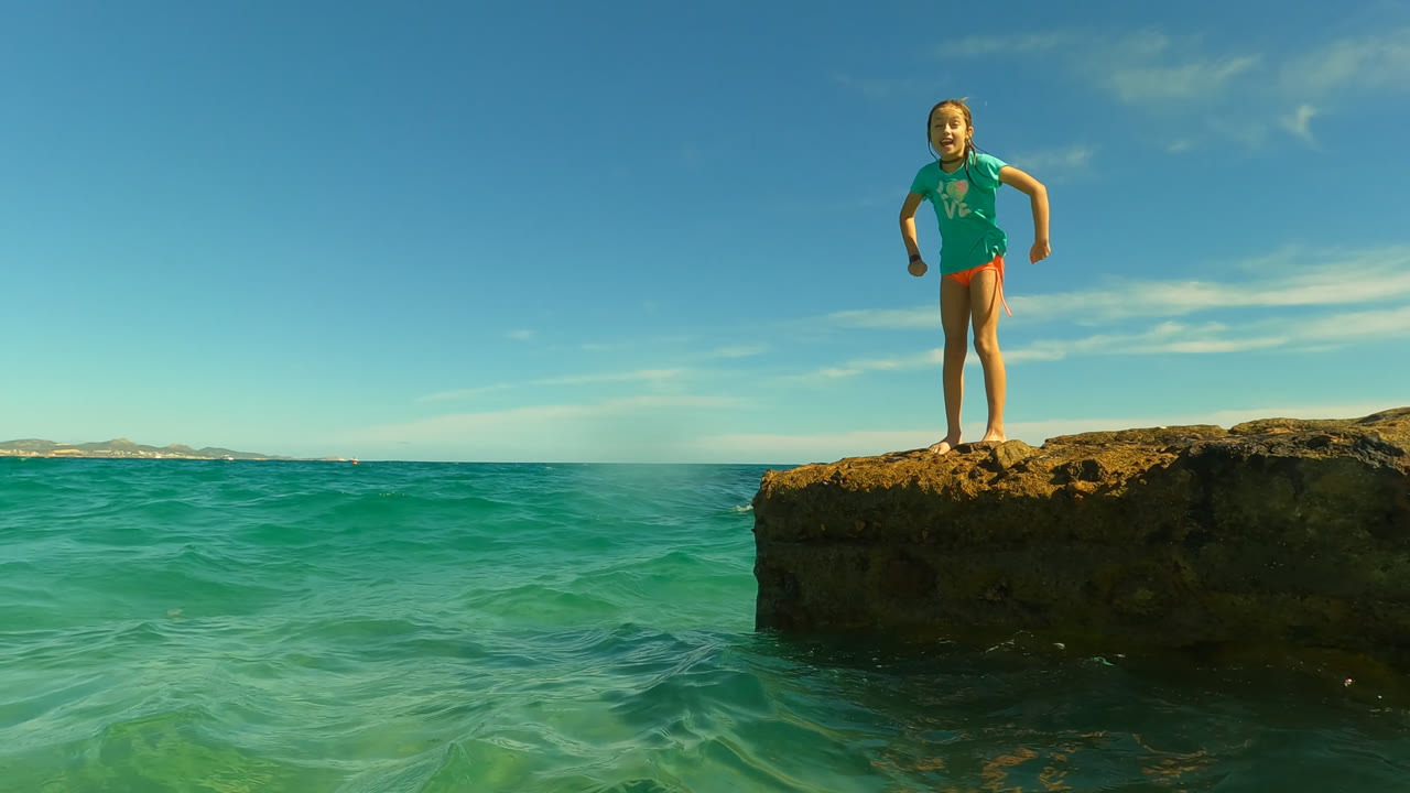 Young girl standing on a rocky ocean ledge under a blue sky.