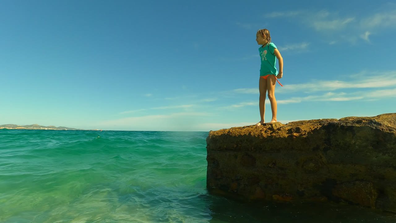 The girl stands near the edge of the rock in San Jose Del Cabo, Mexico, looking down toward the water as small waves move below the ledge.