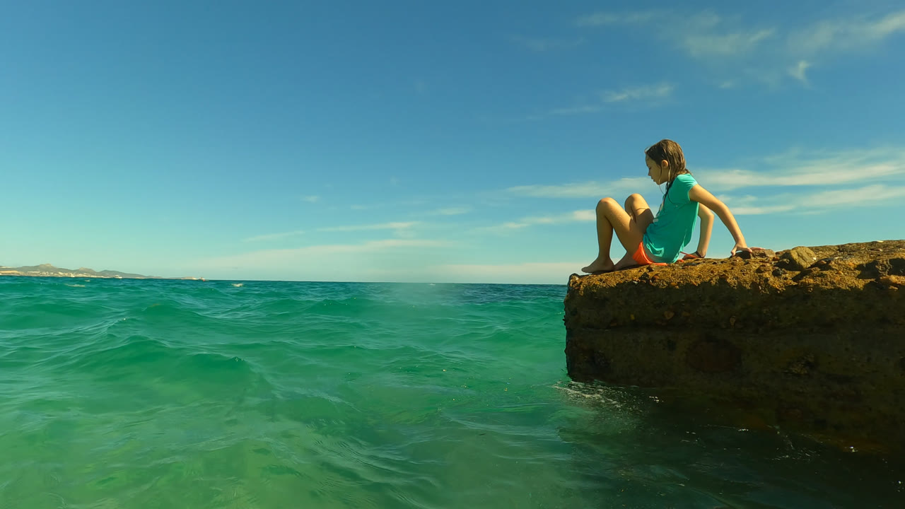 A young girl sits on a rough rock ledge at Palmilla in San Jose Del Cabo, Mexico, with shallow green-blue water filling the foreground and a clear horizon behind her.