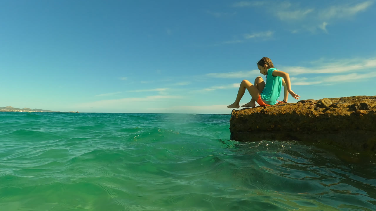 The girl leans slightly forward on the rock at Palmilla in San Jose Del Cabo, Mexico, while rippled shallow water spreads across the lower half of the frame.
