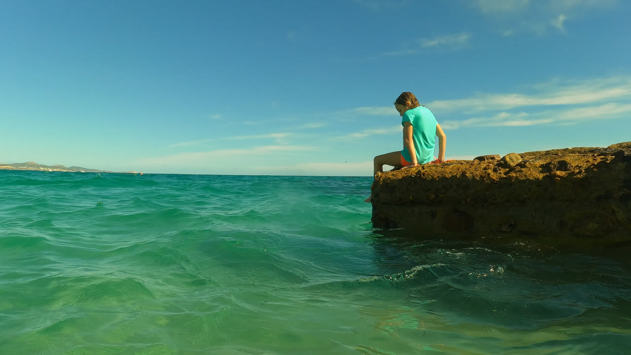 Girl seated near the right edge of a rock over clear ocean