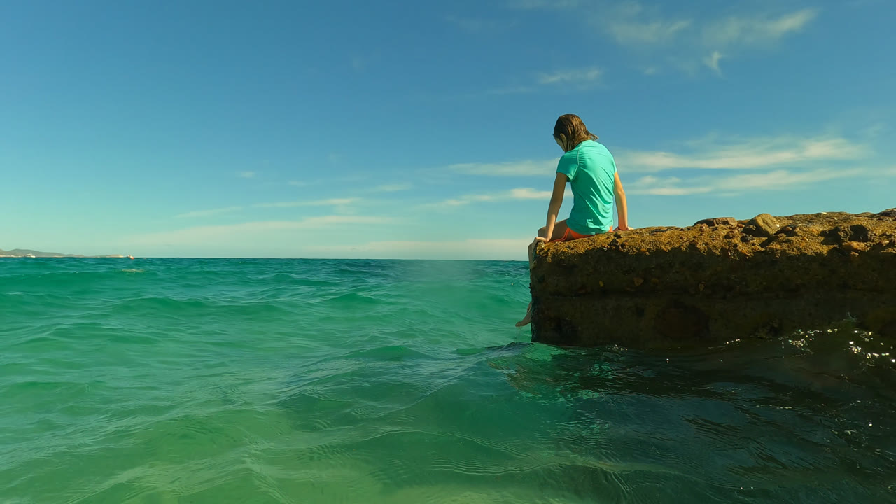 Girl sitting on a rocky ledge above turquoise ocean water.