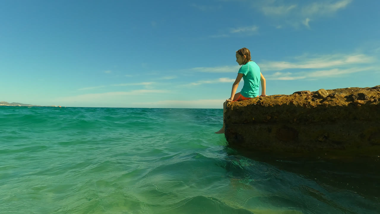At San Jose Del Cabo, Mexico, the seated girl remains on the rock while small ripples darken the water near the ledge and a thin cloud band crosses the blue sky.