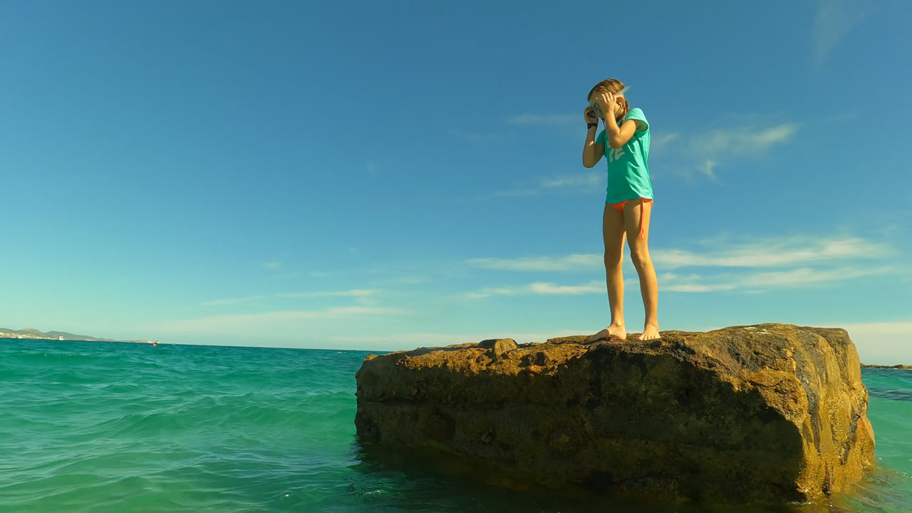 At 0:00 in San Jose Del Cabo, Mexico, a young girl stands on a low rock above turquoise water with her hands near her face and a clear blue sky behind her.