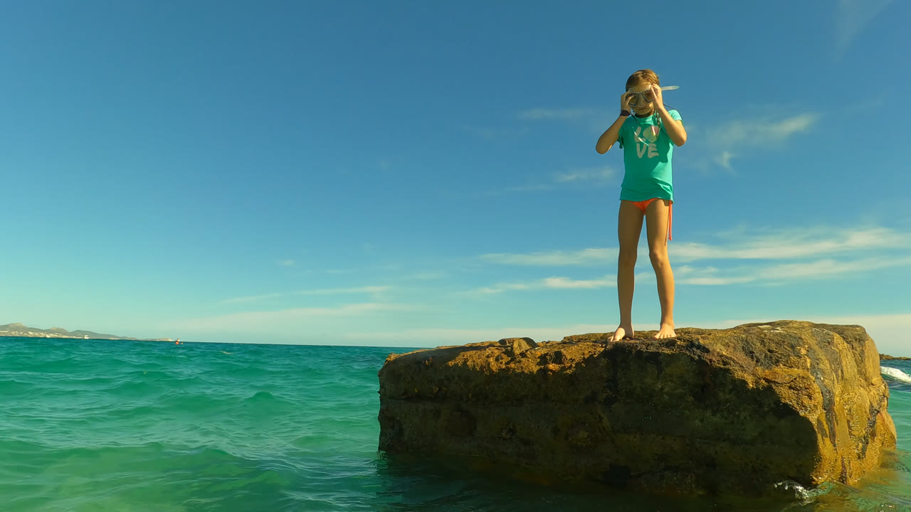 At 0:01 in San Jose Del Cabo, Mexico, the girl faces the camera from the center of the rock while turquoise waves fill the foreground and a distant shoreline sits on the horizon.