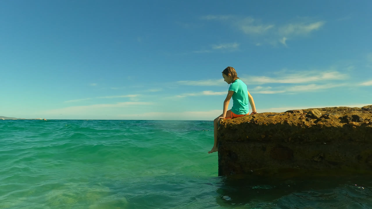 A young girl sits near the left side of a rough ocean rock in San Jose Del Cabo, Mexico, with turquoise water filling the foreground and a blue sky behind her.