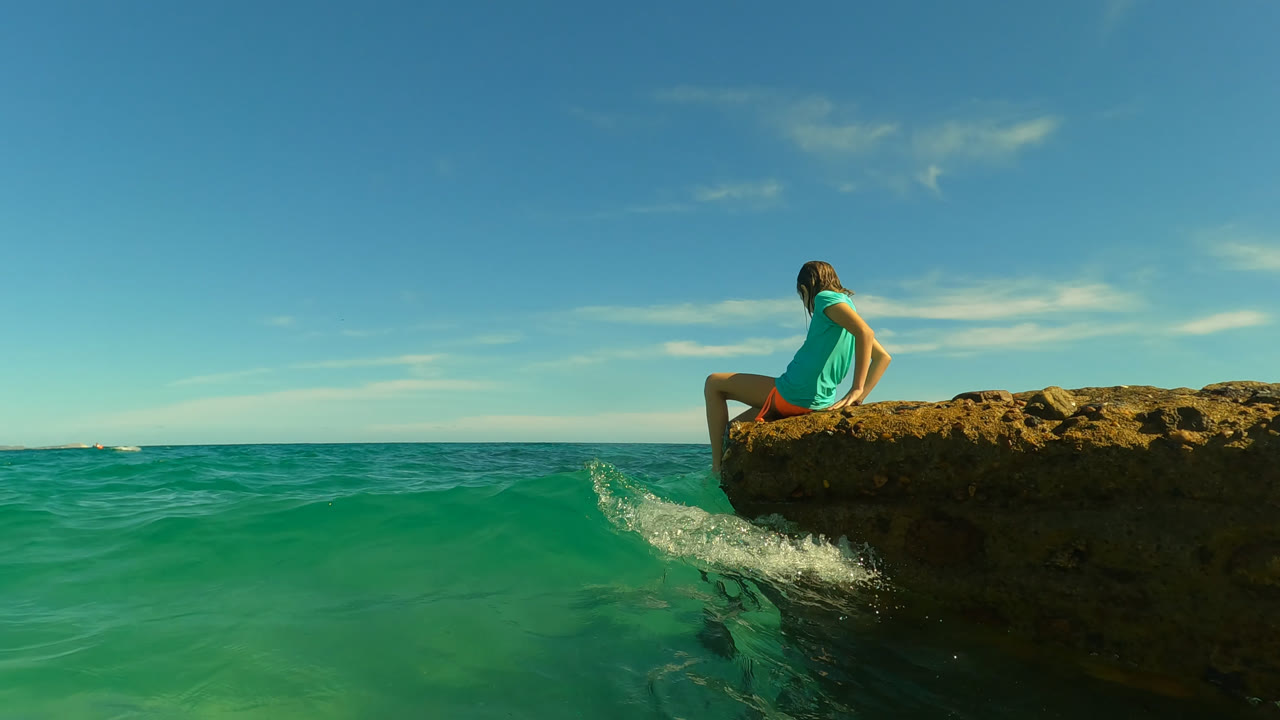 The girl sits on the rock ledge in San Jose Del Cabo, Mexico, while a small splash of water rises against the rock below her.