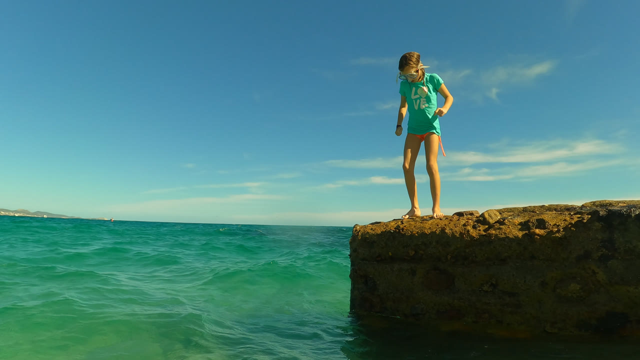Young girl standing on a rocky ledge above turquoise ocean water.