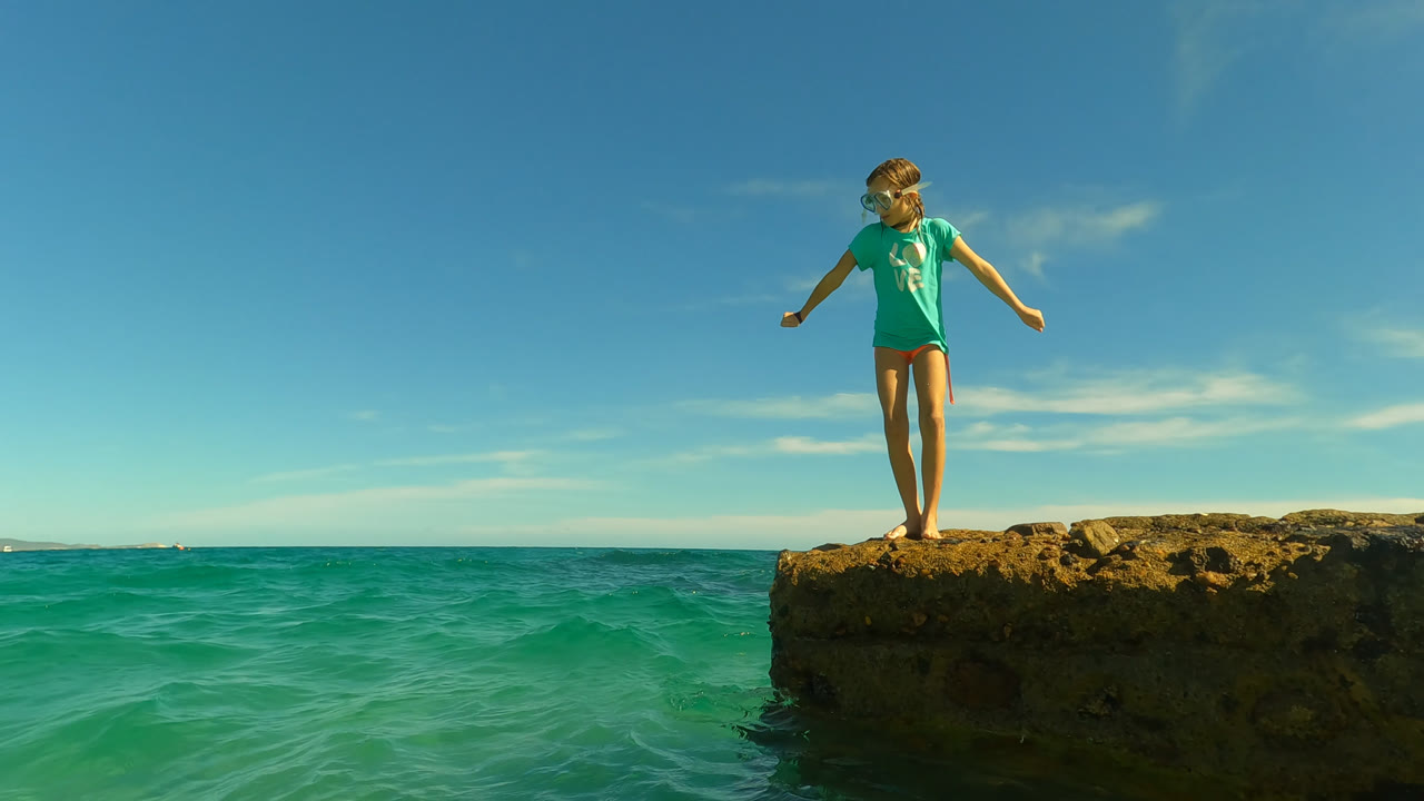 At 0:01 in San Jose Del Cabo, Mexico, the girl holds her arms slightly away from her sides while standing on the rock ledge above rippled turquoise water.