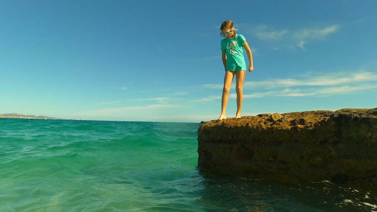 Child standing on a rocky ledge above turquoise ocean water.