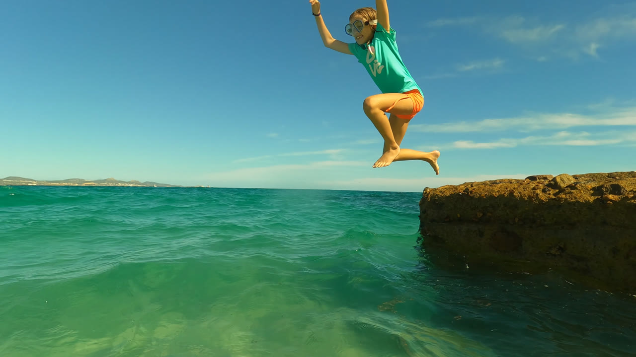The child is airborne over turquoise water between rough rock ledges in San Jose Del Cabo, Mexico, with one arm raised and the horizon behind.