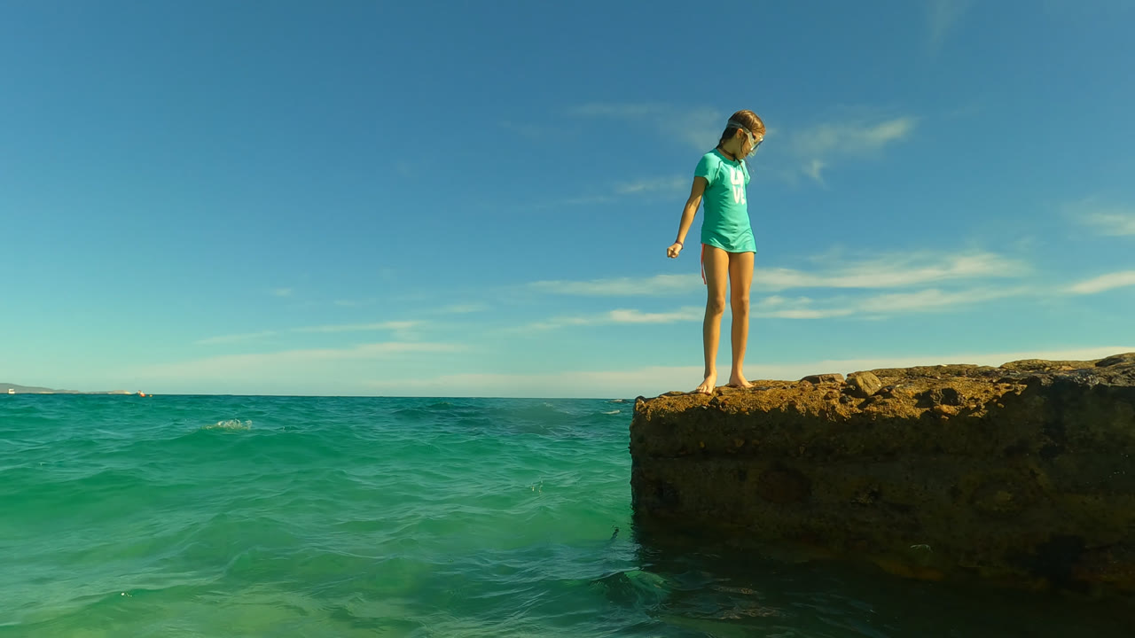 At Palmilla in San Jose Del Cabo, Mexico, a young girl stands near the edge of a rough rock ledge above green-blue ocean water, looking down as the horizon stretches behind her.