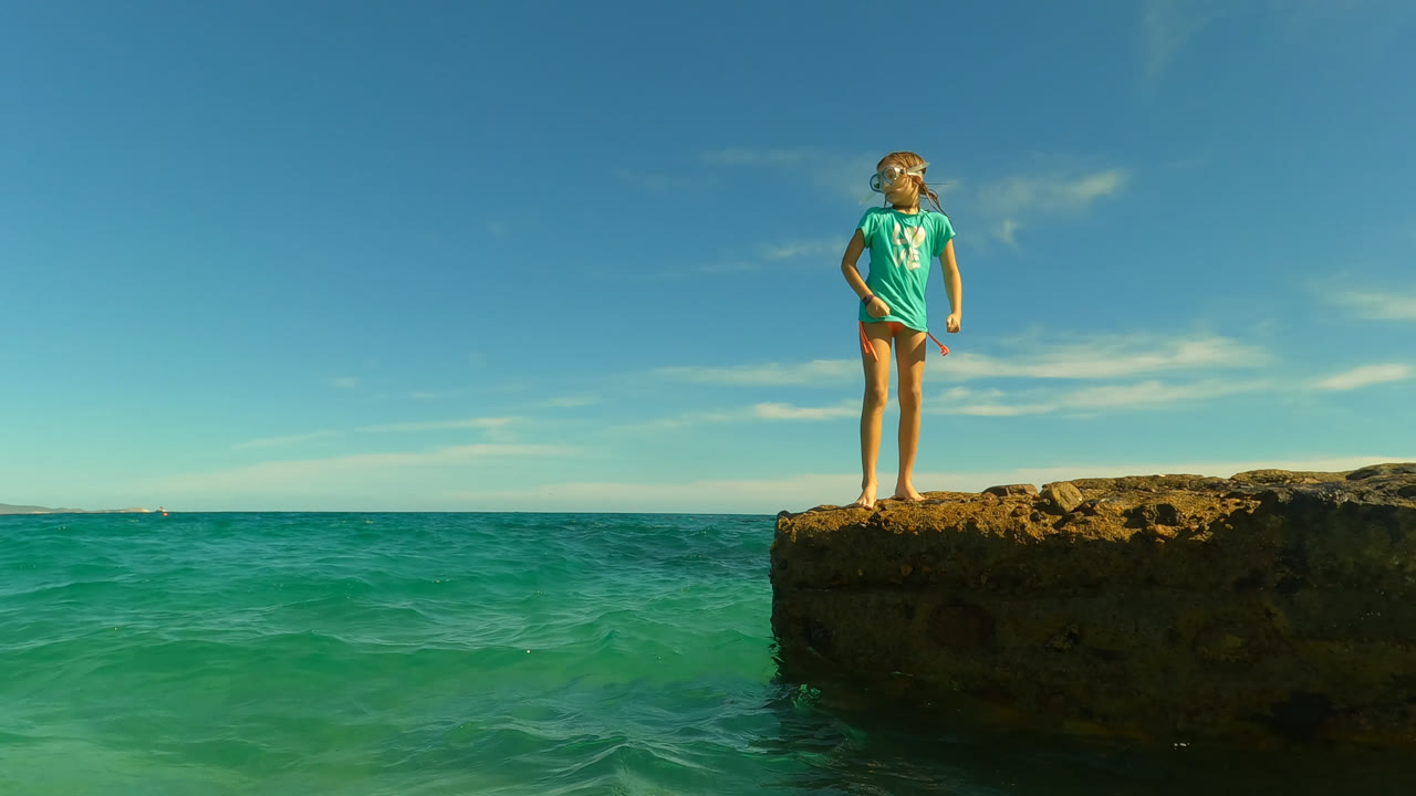 The girl remains on the rocky outcrop at Palmilla, turned slightly to one side with sunlit water, a low coastline, and thin clouds visible across the background.