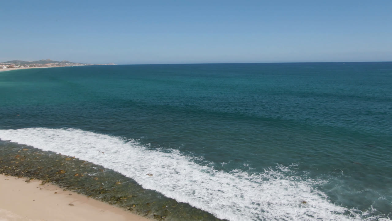 Aerial view over Costa Azul Los Cabos with blue ocean, white surf along a rocky nearshore strip, pale sand at the lower edge, distant coastline, and no visible people.