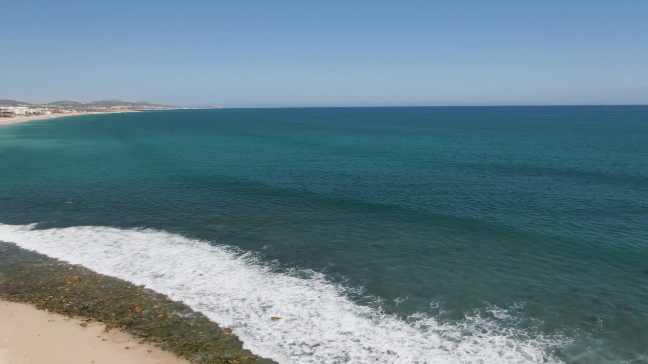 Drone-height view of Costa Azul Los Cabos with turquoise water across the frame, a foamy wave line crossing the lower left, sandy beach at the bottom edge, and no visible people.