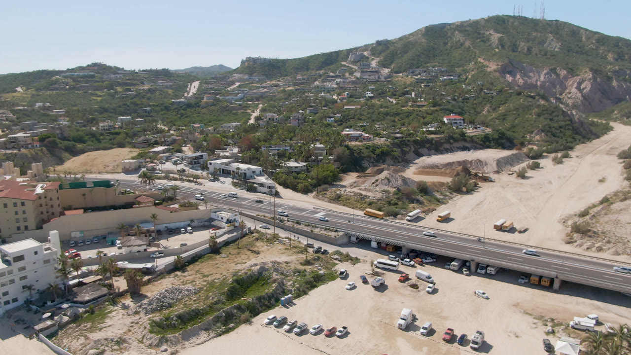 Aerial view in Costa Azul Los Cabos with the bridge running across the lower frame, moving road traffic, parked vehicles below, sandy graded land, hillside roads, and no visible people.