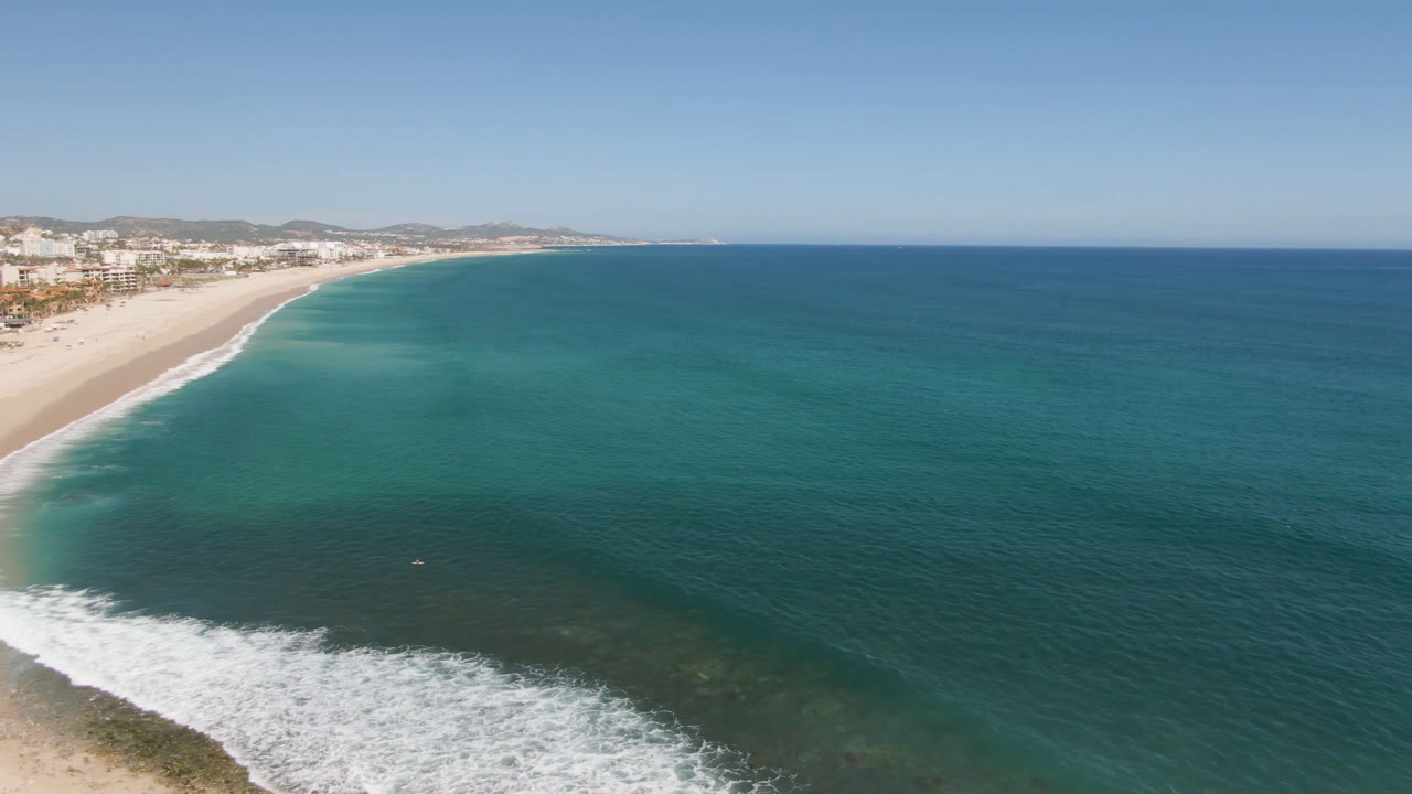 Opening aerial frame over Costa Azul in Los Cabos, with white sand beach at left, blue ocean filling the frame, low surf, distant buildings, and no visible people.