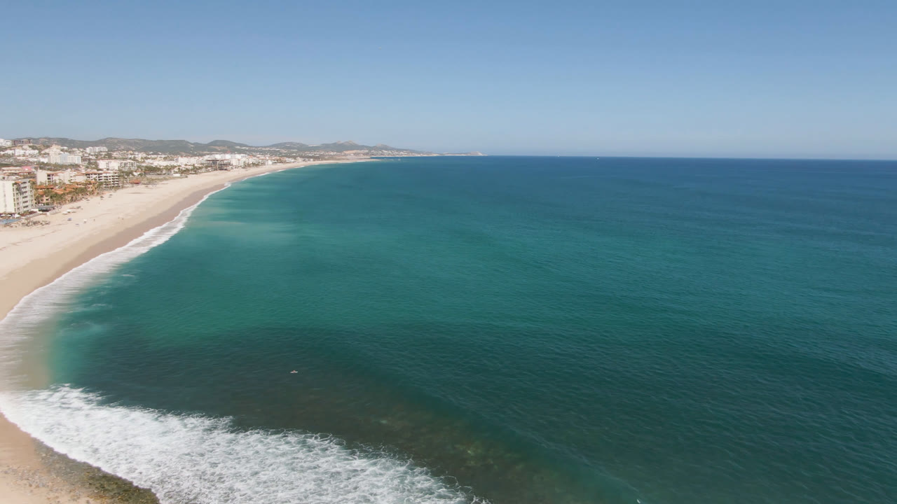 Aerial frame over Costa Azul showing the shoreline sweeping from lower left into the distance, with turquoise shallows, darker offshore water, beachfront buildings, and no visible people.