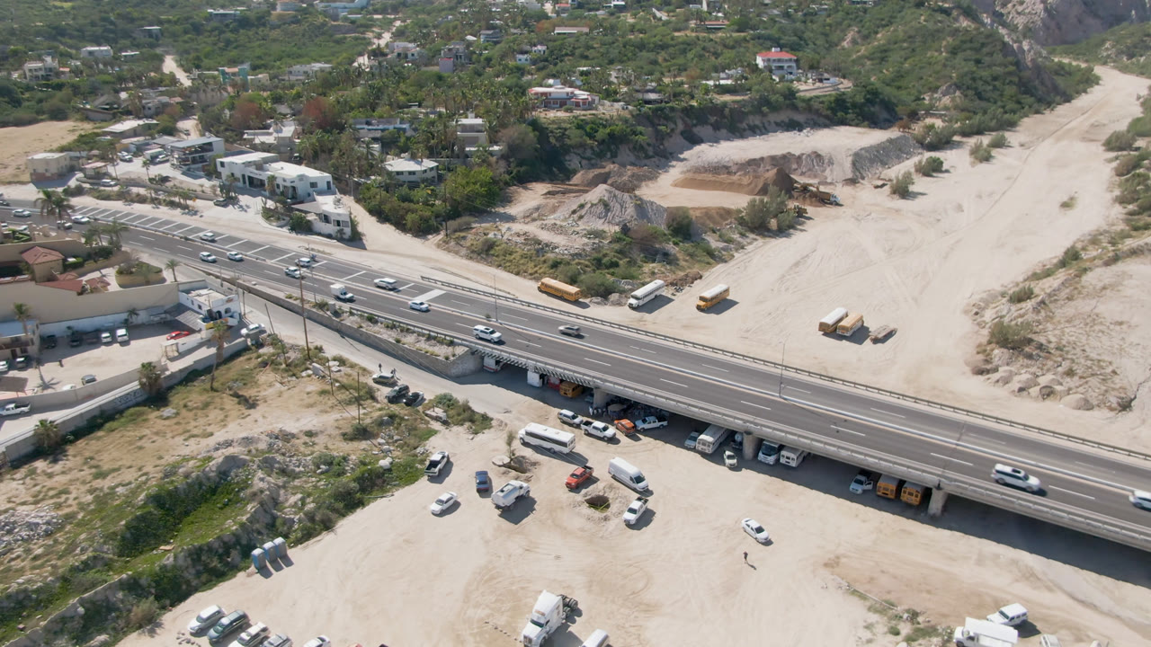 A high-angle aerial frame in Costa Azul, Los Cabos, shows a highway bridge crossing pale sandy ground with moving cars, buses, parked vehicles, hillside homes, and no clearly visible people.