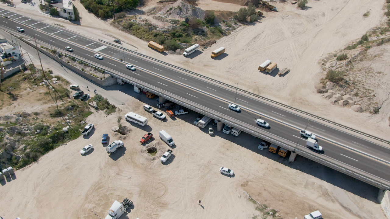Opening overhead frame of a highway bridge in Costa Azul Los Cabos, Baja California Sur, with cars on the road, parked vehicles below, sparse shrubs, and no clearly visible people.