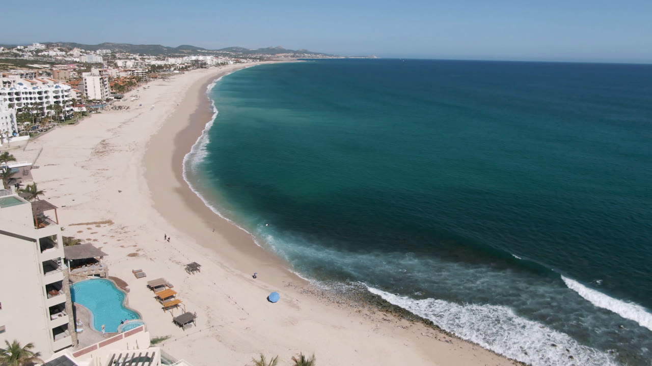 Elevated view over Costa Azul Los Cabos with a turquoise pool at lower left, beachfront hotel buildings, a broad sandy beach, blue ocean, white surf, distant hills, and tiny people visible on the sand.