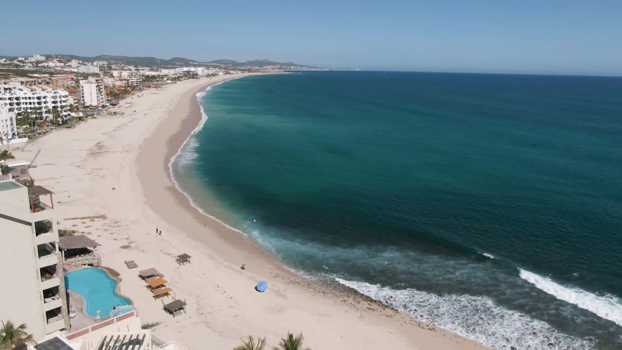 At 0:01, Costa Azul Los Cabos appears from an elevated angle with hotel balconies near the foreground, a pool below, pale sand curving along the shore, blue-green water, and small people visible on the beach.