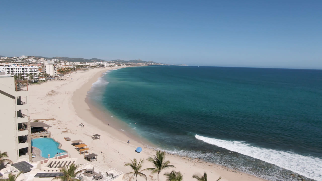 Opening frame of Costa Azul Los Cabos beach with a white hotel edge on the left, turquoise shallows, darker offshore water, palms at the bottom, and no visible people.