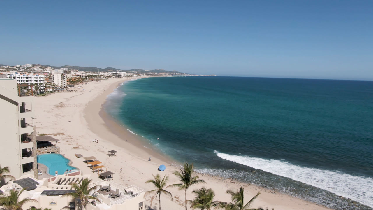 Second frame shows Costa Azul Los Cabos shoreline with the hotel facade tight on the left, a pool below, small beach setups on the sand, and no visible people.