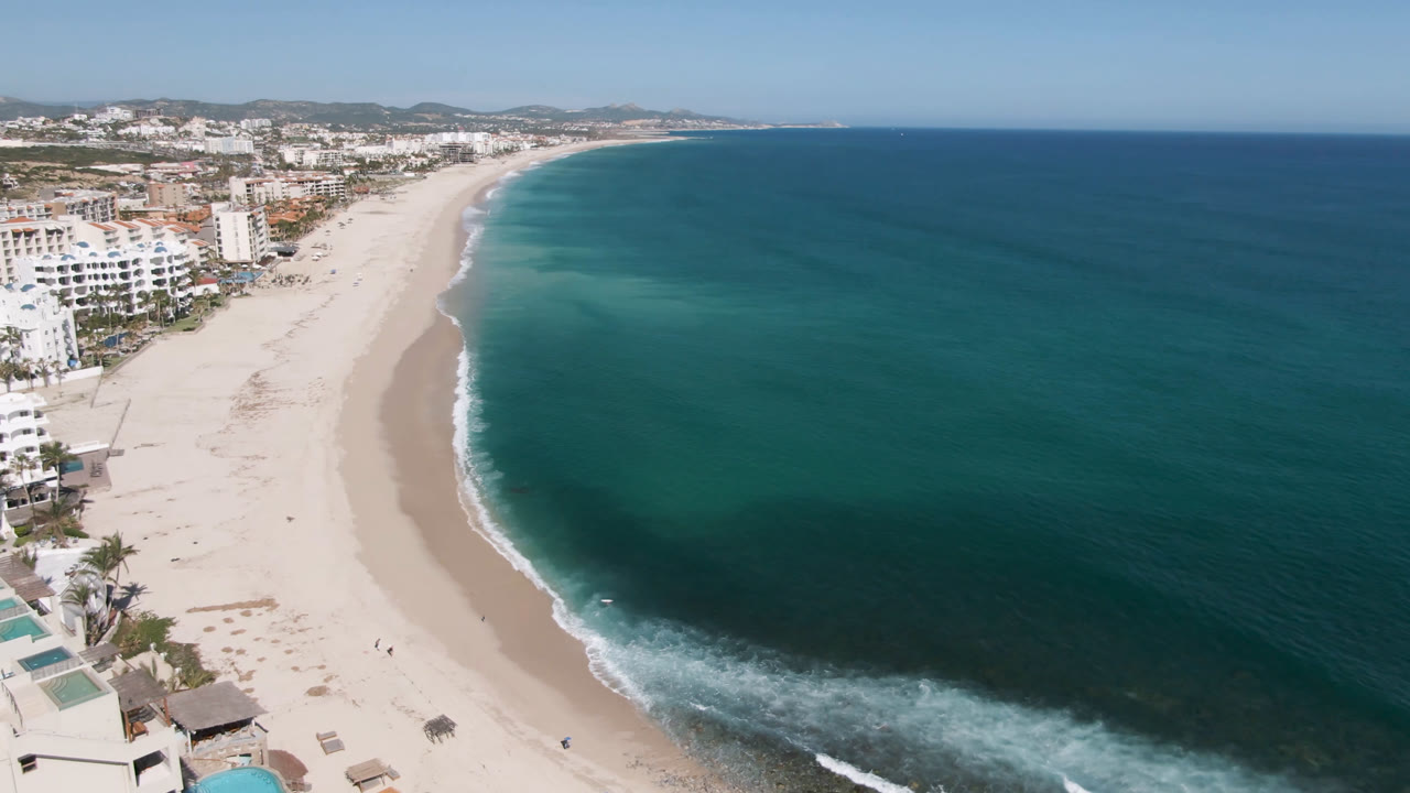 Aerial view of a curving white sand beach beside blue ocean and beachfront buildings.