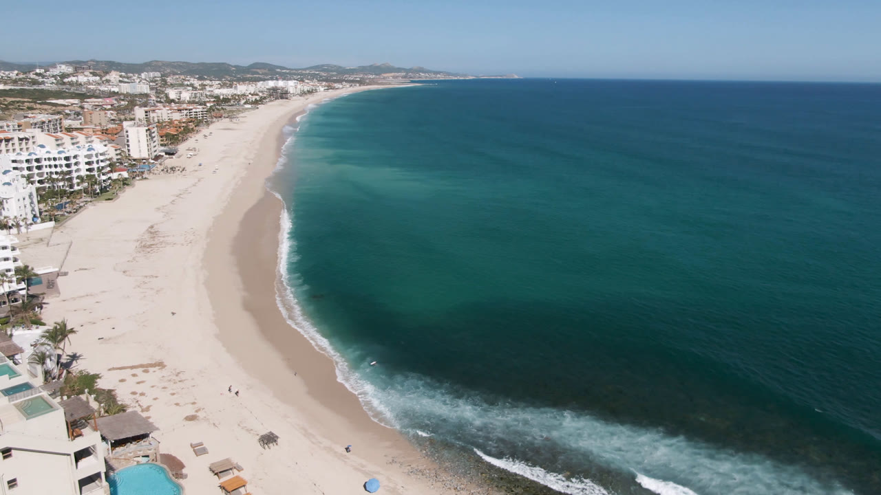 At one second, the Costa Azul Los Cabos shoreline fills the frame with pale sand, a narrow foam line, turquoise shallows, ocean water to the right, and no clearly visible people.