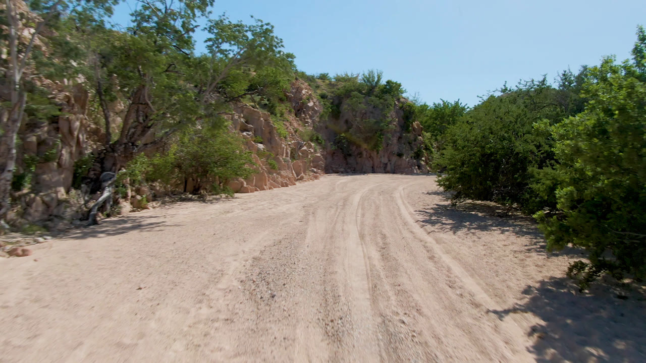 Opening frame of a pale dirt road in Cabo Pulmo, Baja California Sur, with tire tracks leading toward rocky cliffs and dense green brush, no visible people.