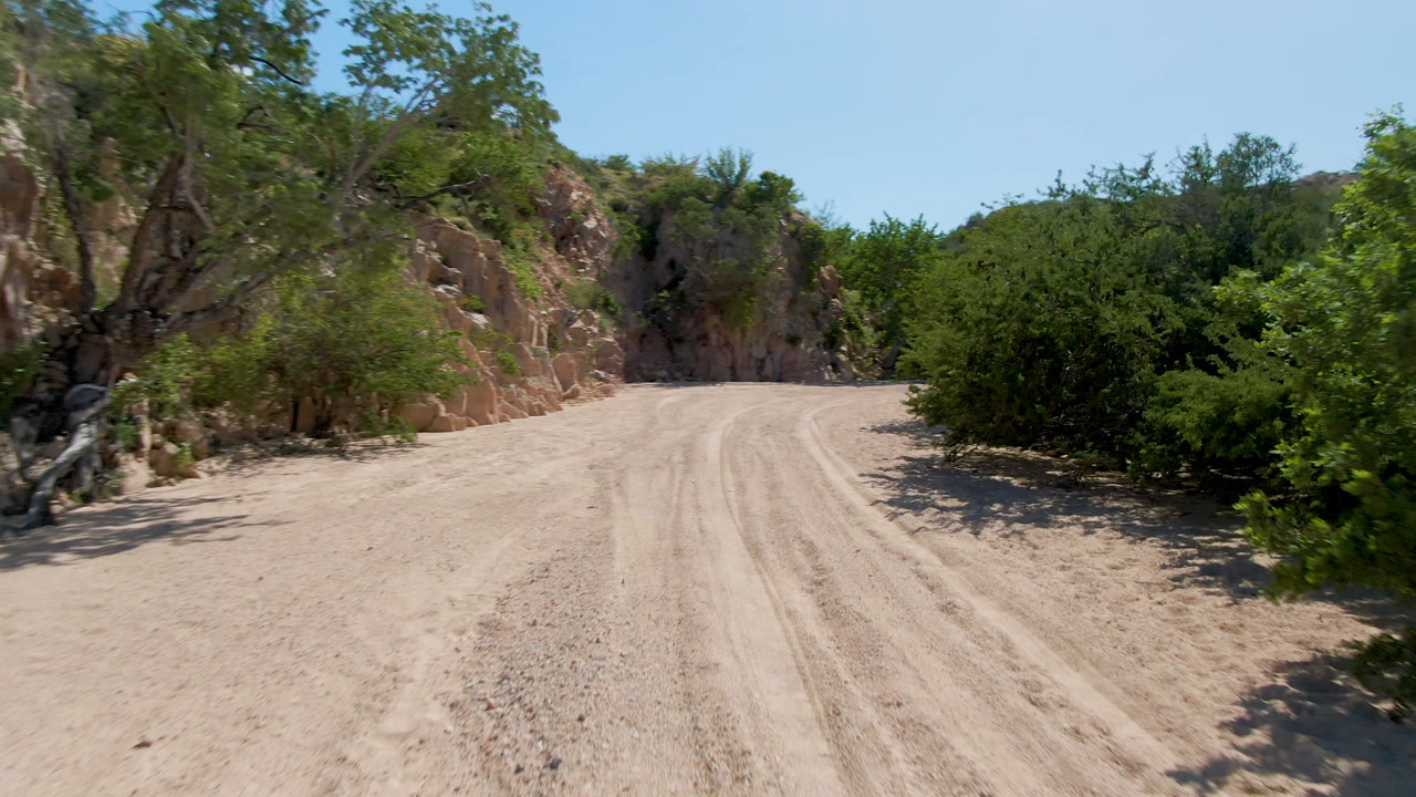A forward road view in Cabo Pulmo shows the sandy track widening in the foreground, shrubs along the right edge, tan rock faces ahead, and no visible people.