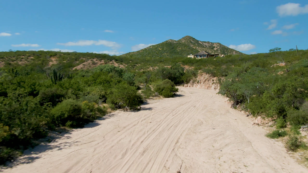 At 0:01, the Cabo Pulmo road remains centered between dense green brush, with the hill and a small distant structure visible beyond it and no visible people.