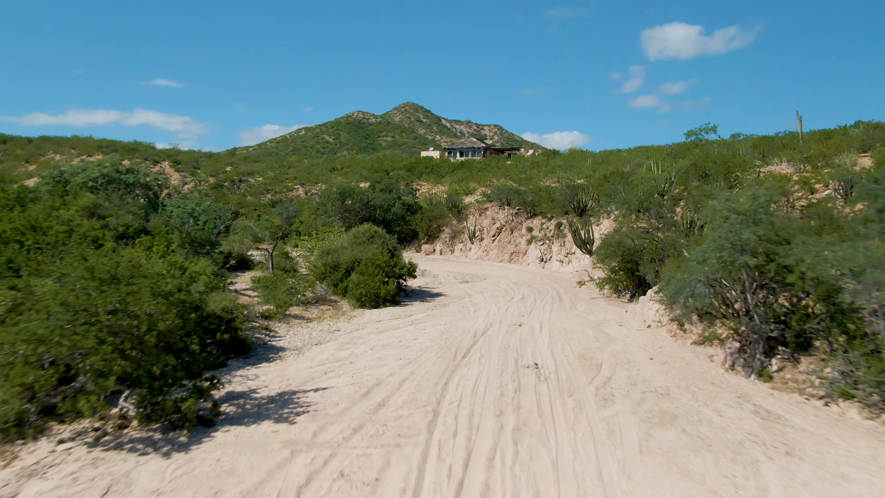 Sandy dirt road through desert brush with a distant house on a hill.