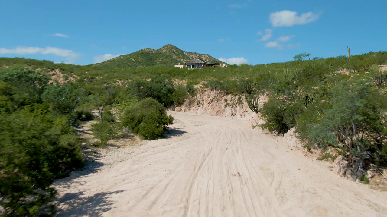 Cabo Pulmo dirt road curving between dense green shrubs and cactus, with a rocky cut bank, a distant hillside house, scattered clouds, and no visible people.