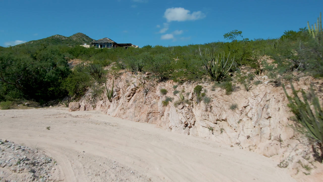 Cabo Pulmo desert roadside view with the road curving through the foreground, rough tan embankment, cactus and green scrub in the midground, a distant hilltop house, and no visible people.