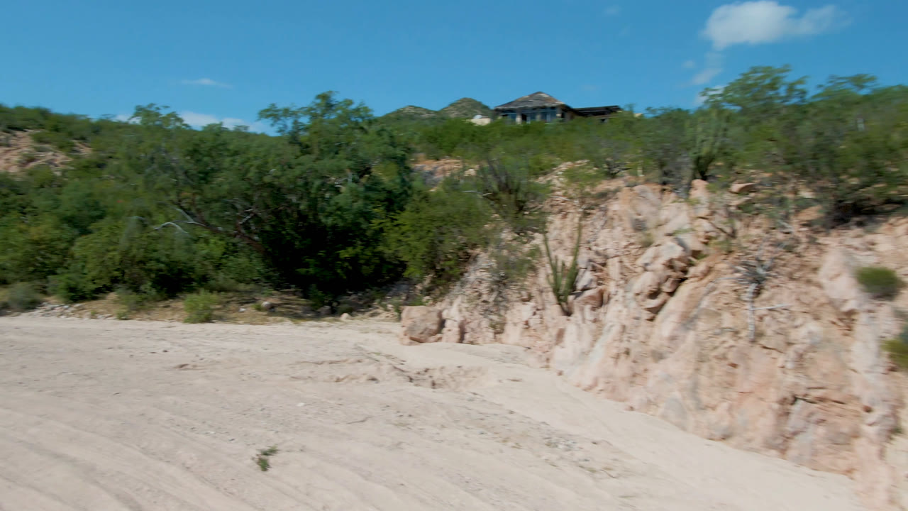 A sandy dirt road runs past a pale eroded rock bank and green desert brush in San Jose Del Cabo, Mexico, with a small distant roofline and no visible people.
