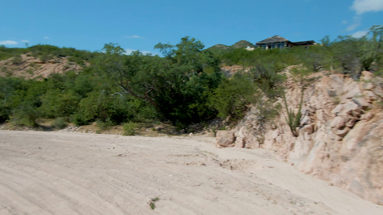 The view continues along the dusty road with pale rock in the foreground, dense green shrubs in the midground, low hills behind them, and no visible people.
