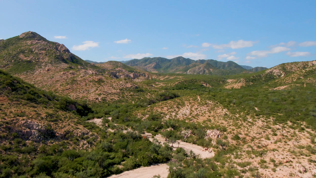 Opening wide aerial view of Cabo Pulmo desert hills, with a pale dirt road in the lower valley, rocky green-brown slopes, distant ridges, blue sky, and no visible people.